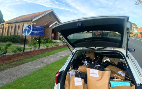 An open car boot containing Christmas Gifts. The car is parked in front of a church signed 'Penrith Presbyterian Church'.